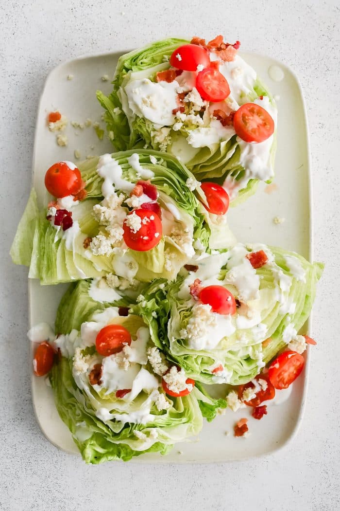 Large white rectangular serving platter with four wedges of iceberg lettuce, cut-side-up, drizzled with homemade blue cheese dressing and sprinkled with crumbled bacon bits, halved cherry tomatoes, and blue cheese crumbles.