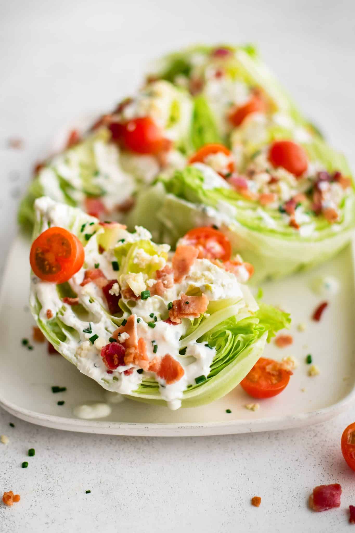 Large white rectangular serving platter with four wedges of iceberg lettuce, cut-side-up, drizzled with homemade blue cheese dressing and sprinkled with crumbled bacon bits, halved cherry tomatoes, chives, and blue cheese crumbles.