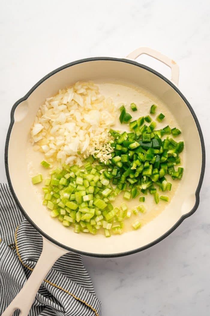 Overhead view of a cream-colored skillet with a black rim and pale handle filled with chopped onion, chopped celery, chopped green bell pepper, and minced garlic in melted butter. The vegetables are separated in small piles before being stirred together. The skillet sits on a light gray marble surface beside a gray-and-white striped kitchen towel with yellow trim.
