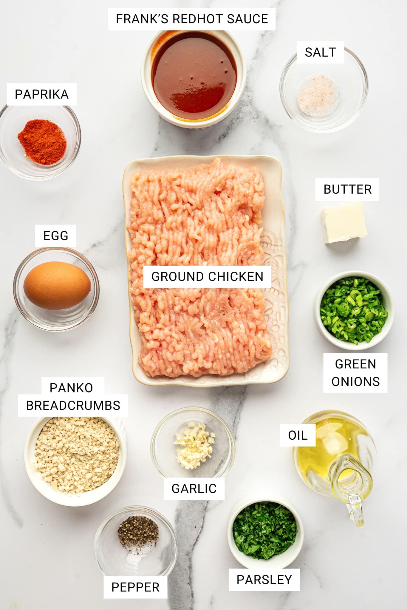 Overhead ingredient layout on a white marble surface with labeled text overlays, including a tray of ground chicken labeled “GROUND CHICKEN,” a bowl of hot sauce labeled “FRANK’S REDHOT SAUCE,” small bowls labeled “PAPRIKA,” “SALT,” “PEPPER,” “EGG,” “PANKO BREADCRUMBS,” “GARLIC,” “PARSLEY,” a small bowl labeled “GREEN ONIONS,” a pat labeled “BUTTER,” and a glass container labeled “OIL.”