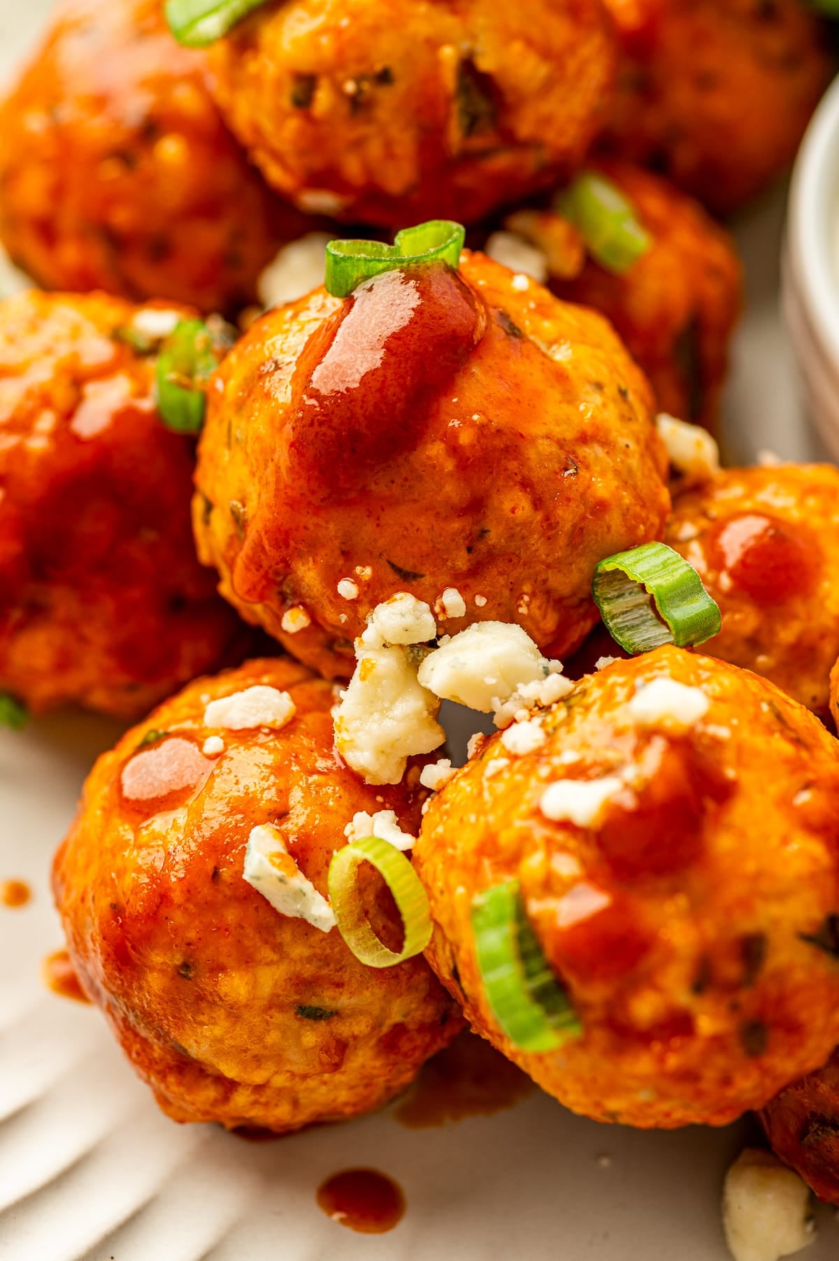 Extreme close-up of glossy buffalo chicken meatballs coated in bright red buffalo sauce, garnished with sliced green onions and crumbled blue cheese, on an off-white ridged plate with a small white ramekin of creamy dip partially visible in the background.