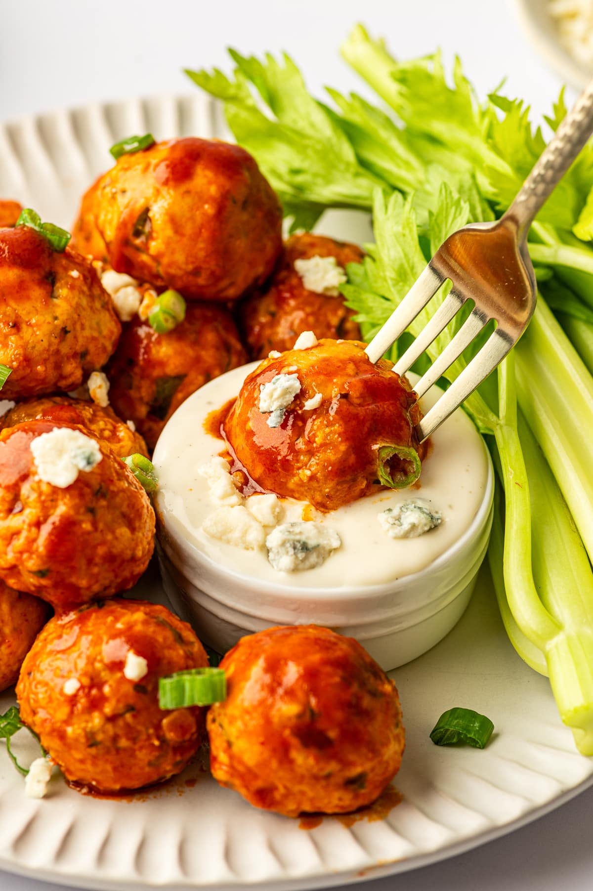 Buffalo chicken meatballs coated in red buffalo sauce on an off-white ridged plate with celery sticks, with a fork holding a meatball dipped into a small white ramekin of creamy dip topped with blue cheese crumbles, plus scattered green onion slices.