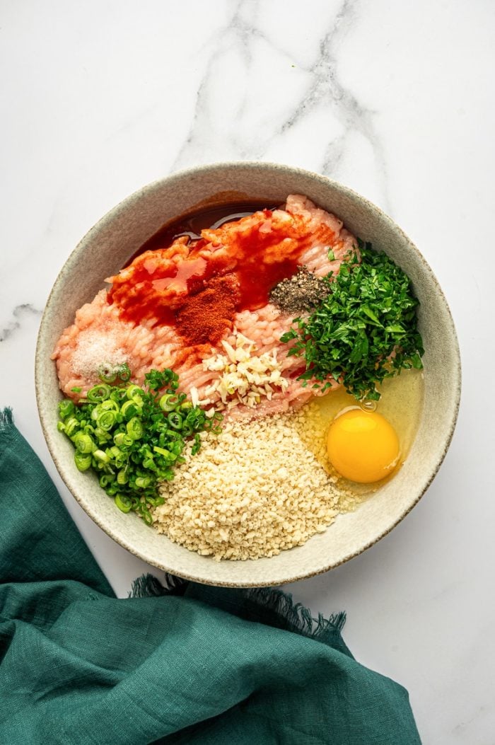 Overhead view of a light gray ceramic bowl filled with buffalo chicken meatball ingredients on a white marble surface, including raw ground chicken, panko breadcrumbs, a whole raw egg, chopped green onions, chopped parsley, minced garlic, paprika, salt, black pepper, and red hot sauce, with a teal green cloth napkin on the right.