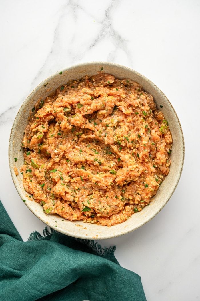 Overhead view of a light gray ceramic bowl holding mixed buffalo chicken meatball mixture, showing ground chicken combined with panko, herbs, and green onions on a white marble surface, with a teal green cloth napkin to the right.