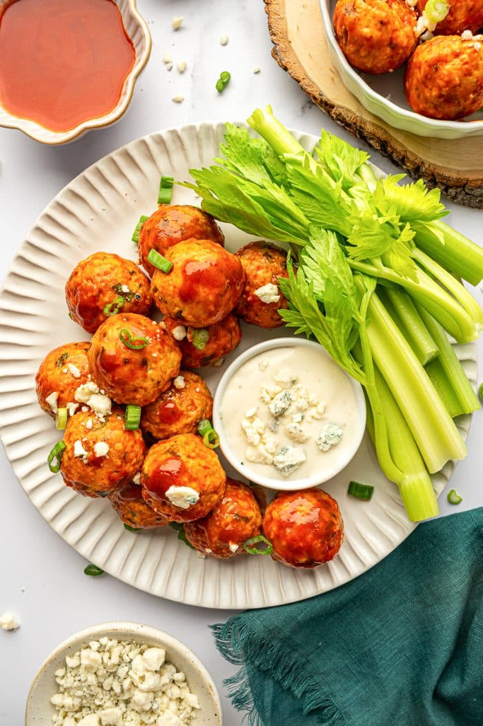 Overhead close view of buffalo chicken meatballs on an off-white ridged plate beside celery sticks and a white ramekin of creamy dip topped with blue cheese crumbles, with a bowl of buffalo sauce partially visible in the top left.