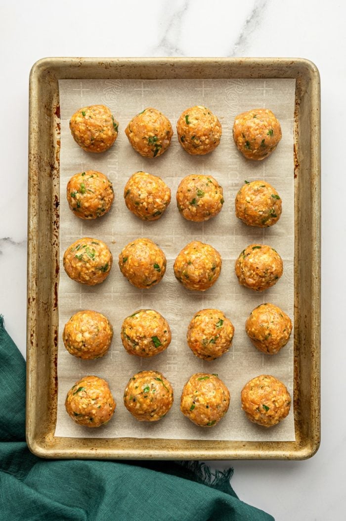 Overhead view of raw buffalo chicken meatballs arranged on a parchment-lined metal baking sheet, spaced apart in neat rows, with a teal green cloth napkin on the right and no text overlay.