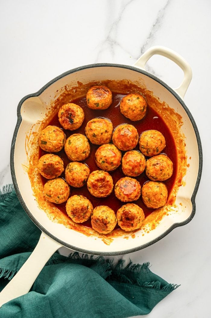 Overhead view of buffalo chicken meatballs coated in red buffalo sauce in a cream enameled skillet, with sauce splashes around the sides, on a white marble surface with a teal green cloth napkin on the right.