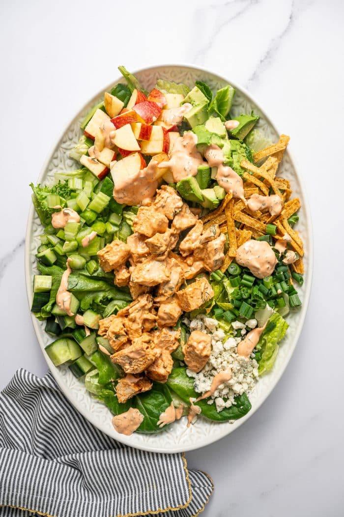 Buffalo chicken salad on a light green oval platter with chopped greens, cucumber, celery, diced apple, avocado, tortilla strips, green onions, blue cheese crumbles, buffalo chicken, and creamy buffalo dressing drizzled over the top on a white marble surface with a black-and-white striped kitchen towel with gold trim in the bottom left corner.