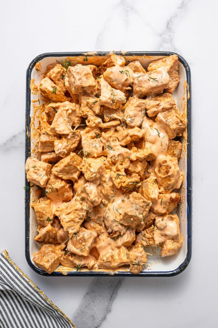 Chopped chicken coated in creamy buffalo dressing with fresh dill in a white rectangular baking dish with a dark rim on a white marble surface with a black-and-white striped kitchen towel with gold trim in the bottom left corner.