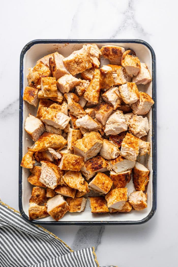 Bite-sized pieces of cooked buffalo chicken salad chicken in a white rectangular baking dish with a dark rim on a white marble surface with a black-and-white striped kitchen towel with gold trim in the bottom left corner.