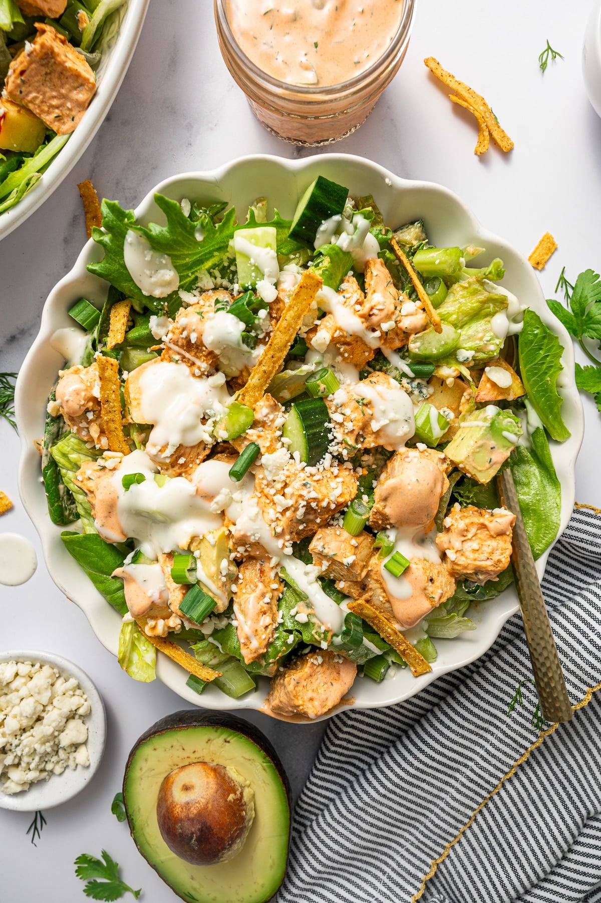 Close-up of buffalo chicken salad in a pale green scalloped bowl with chopped lettuce, buffalo chicken, cucumber, avocado, apple, tortilla strips, green onions, blue cheese crumbles, creamy buffalo dressing, and ranch dressing, with a spoon in the bowl, a jar of buffalo dressing, half an avocado, and a small dish of blue cheese crumbles on a white marble surface with a black-and-white striped kitchen towel.