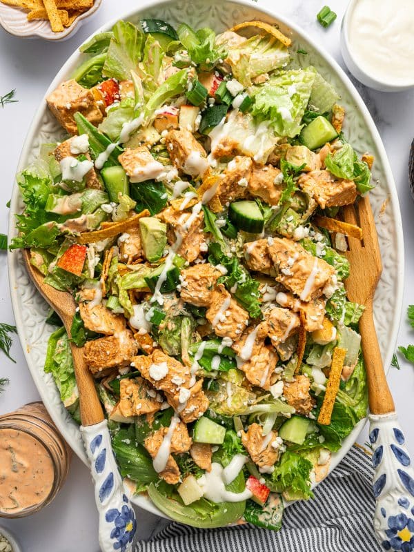 Close-up of tossed buffalo chicken salad on a light green oval platter with chopped lettuce, cucumber, avocado, apple, tortilla strips, green onions, buffalo chicken, blue cheese crumbles, creamy buffalo dressing, and ranch dressing, with wooden serving spoons, a bowl of ranch, and a jar of buffalo dressing partially visible on a white marble surface.