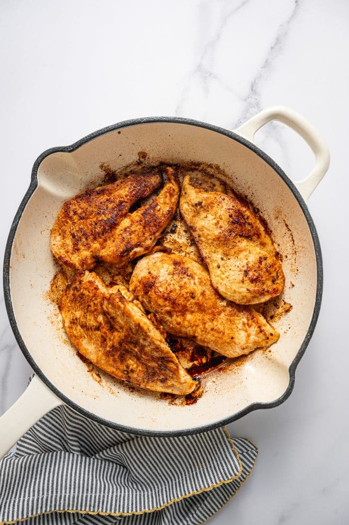 Cooked chicken breasts with golden brown seasoning in a cream-colored skillet with a black rim on a white marble surface with a black-and-white striped kitchen towel with gold trim in the bottom left corner.