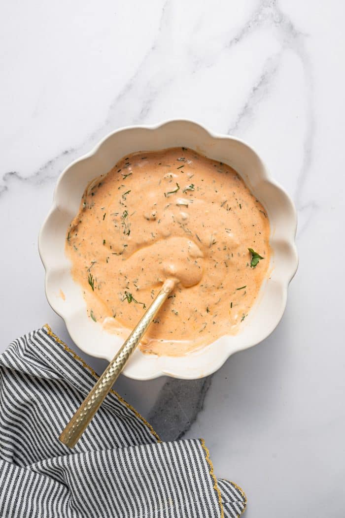 Creamy buffalo dressing with visible fresh dill and herbs in a white scalloped bowl with a gold spoon on a white marble surface with a black-and-white striped kitchen towel with gold trim in the bottom left corner.