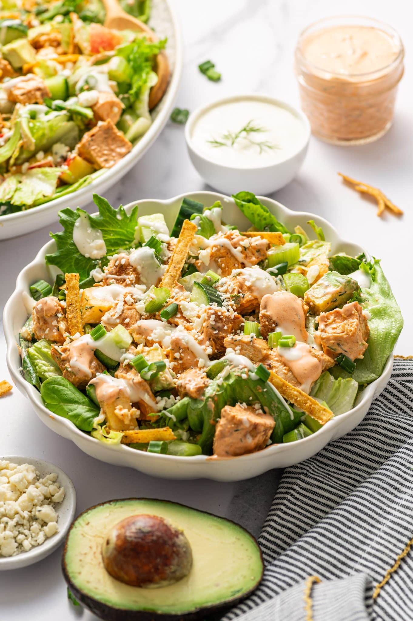 Individual serving of buffalo chicken salad in a pale green scalloped bowl filled with chopped greens, buffalo chicken, cucumber, avocado, tortilla strips, green onions, and crumbled blue cheese, drizzled with creamy orange buffalo dressing and white ranch dressing. In the background, a larger serving platter of salad is softly blurred, along with a small white bowl of ranch dressing topped with dill and a glass jar of buffalo dressing. A halved avocado, a small white dish of blue cheese crumbles, and a black-and-white striped kitchen towel with gold trim sit on the white marble surface.