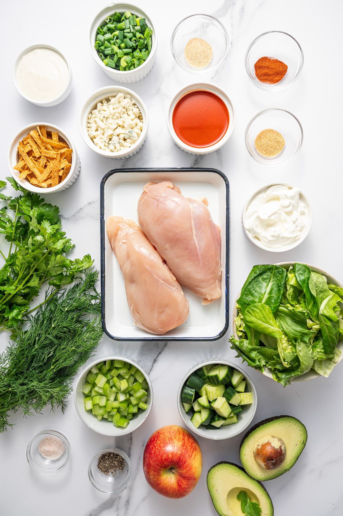 Overhead view of buffalo chicken salad ingredients arranged on a white marble surface. Two raw chicken breasts sit in a white rectangular baking dish with a dark rim, surrounded by small white bowls of ranch dressing, chopped green onions, crumbled blue cheese, buffalo sauce, sour cream, chopped celery, chopped cucumber, tortilla strips, and mixed greens. Fresh cilantro and dill are arranged on the left side, with small glass bowls of salt, black pepper, onion powder, garlic powder, and paprika near the chicken. A whole red apple and two avocado halves sit at the bottom right.