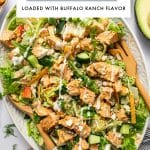 Vertical Pinterest graphic showing a large platter of buffalo chicken salad on a pale green oval platter with wooden serving spoons, chopped greens, buffalo chicken, cucumber, apple, avocado, tortilla strips, blue cheese crumbles, and drizzles of creamy orange buffalo dressing and white ranch dressing. At the top, a large white text box overlay reads โBUFFALO CHICKEN SALADโ in black letters, with a thin gray line underneath and smaller black text below that reads โLOADED WITH BUFFALO RANCH FLAVOR.โ Near the bottom, small black text reads โthe forked spoon.โ A jar of buffalo dressing appears at the lower left, and scattered dill, tortilla strips, and avocado are visible around the platter.