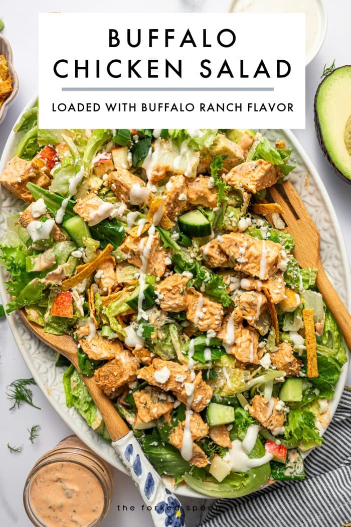 Vertical Pinterest graphic showing a large platter of buffalo chicken salad on a pale green oval platter with wooden serving spoons, chopped greens, buffalo chicken, cucumber, apple, avocado, tortilla strips, blue cheese crumbles, and drizzles of creamy orange buffalo dressing and white ranch dressing. At the top, a large white text box overlay reads “BUFFALO CHICKEN SALAD” in black letters, with a thin gray line underneath and smaller black text below that reads “LOADED WITH BUFFALO RANCH FLAVOR.” Near the bottom, small black text reads “the forked spoon.” A jar of buffalo dressing appears at the lower left, and scattered dill, tortilla strips, and avocado are visible around the platter.
