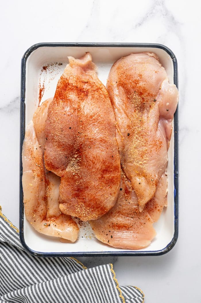 Raw chicken breasts seasoned with paprika, garlic powder, onion powder, salt, and black pepper in a white rectangular baking dish with a dark rim on a white marble surface with a black-and-white striped kitchen towel with gold trim in the bottom left corner.