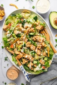 Tossed buffalo chicken salad on a light green oval platter with wooden serving spoons, chopped greens, cucumber, apple, avocado, tortilla strips, blue cheese crumbles, green onions, buffalo chicken, creamy buffalo dressing, and ranch dressing, surrounded by a jar of buffalo dressing, a bowl of ranch, avocado halves, and fresh dill on a white marble surface with a black-and-white striped kitchen towel.