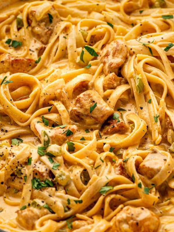 Close-up view of Cajun Chicken Alfredo in a skillet, showing fettuccine coated in creamy orange-tinted Alfredo sauce with bite-sized browned chicken pieces, diced green bell pepper and celery, chopped parsley, and cracked black pepper. The sauce clings to the noodles and chicken.
