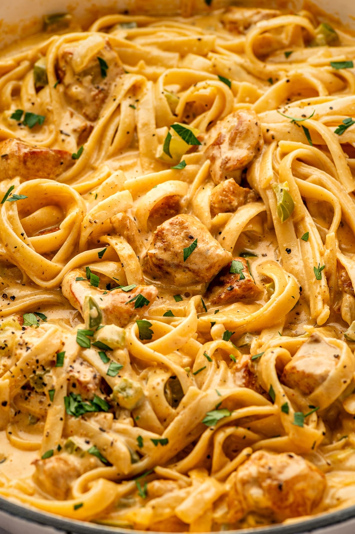 Close-up view of Cajun Chicken Alfredo in a skillet, showing fettuccine coated in creamy orange-tinted Alfredo sauce with bite-sized browned chicken pieces, diced green bell pepper and celery, chopped parsley, and cracked black pepper. The sauce clings to the noodles and chicken.