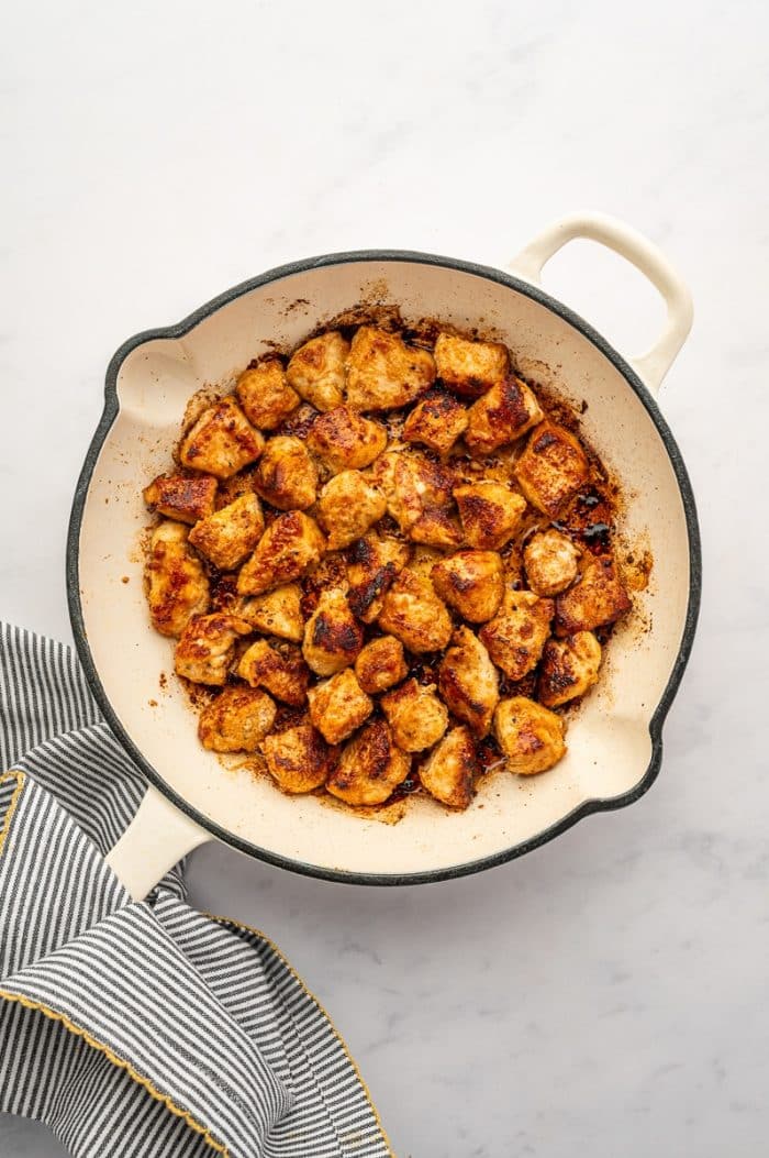 Overhead view of browned bite-sized chicken pieces cooking in a cream-colored skillet with a black rim and pale handle. The chicken is golden brown with darker caramelized edges, and browned bits are visible on the bottom of the skillet. The pan sits on a light gray marble surface beside a gray-and-white striped kitchen towel with yellow trim.