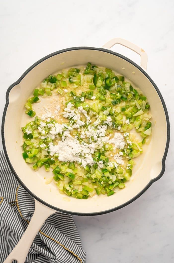 Overhead view of a cream-colored skillet with a black rim and pale handle filled with softened onion, celery, and green bell pepper. White flour is sprinkled over the vegetables before being stirred in. The skillet sits on a light gray marble surface beside a gray-and-white striped kitchen towel with yellow trim.