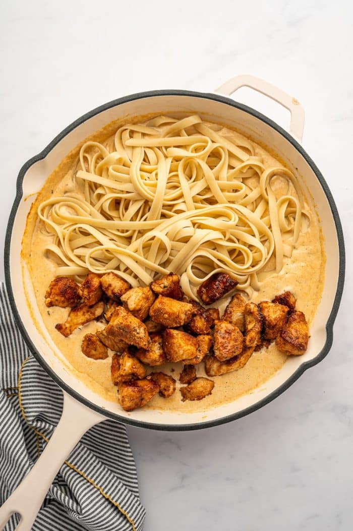 Overhead view of a cream-colored skillet with a black rim and pale handle filled with orange-tinted Cajun Alfredo sauce, cooked fettuccine, and browned bite-sized chicken pieces. The pasta and chicken have been added on top of the sauce before being fully tossed together. The skillet sits on a light gray marble surface beside a gray-and-white striped kitchen towel with yellow trim.