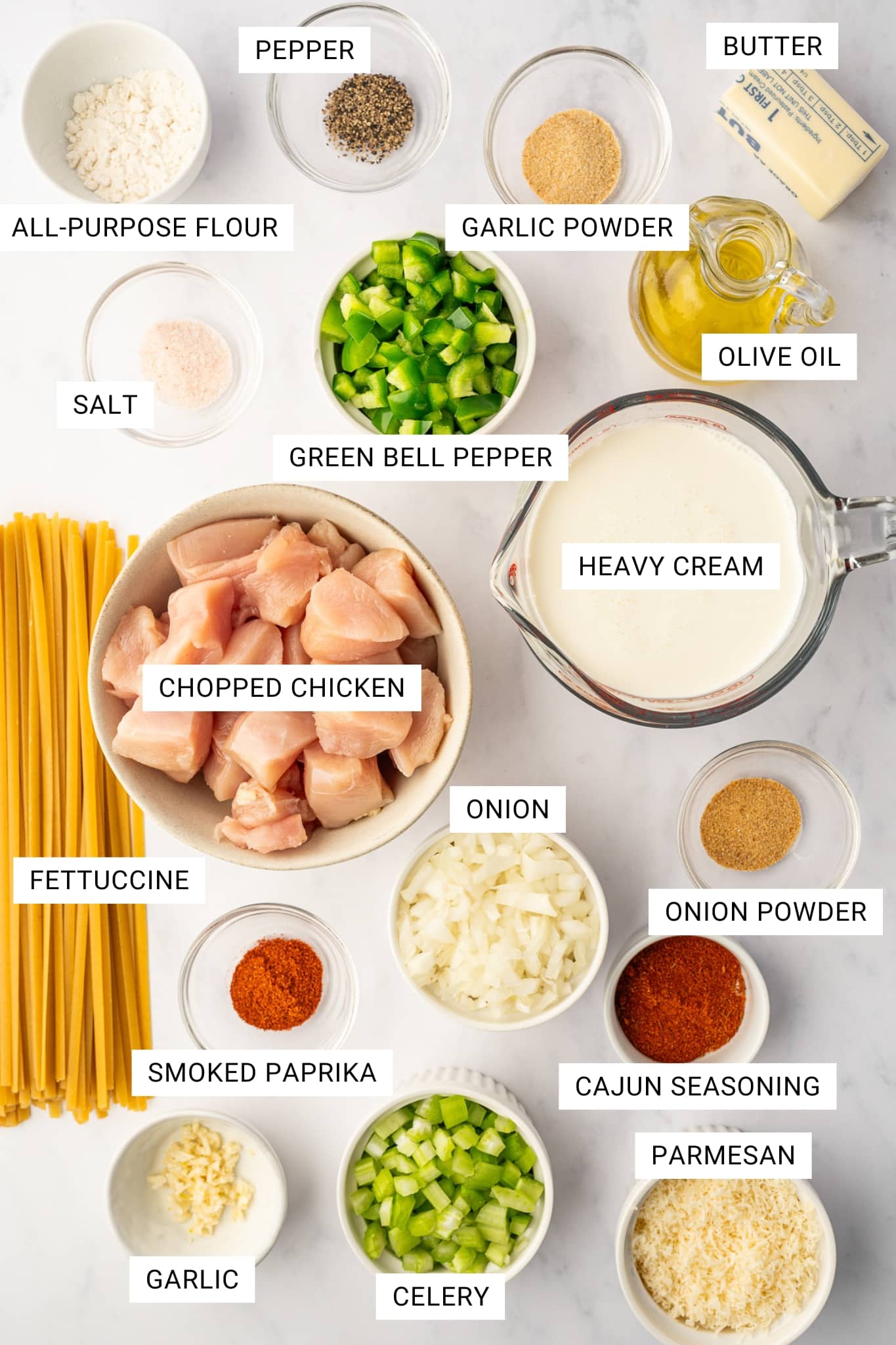 Overhead ingredient photo for Cajun Chicken Alfredo on a light gray marble surface with white text labels placed over each ingredient. Visible ingredients include chopped chicken in a beige bowl, fettuccine, chopped onion, chopped celery, chopped green bell pepper, heavy cream in a glass measuring cup, olive oil in a glass bottle, butter, all-purpose flour, salt, pepper, garlic powder, onion powder, smoked paprika, Cajun seasoning, minced garlic, and grated Parmesan. Each ingredient is labeled with text overlays including CHOPPED CHICKEN, FETTUCCINE, ONION, CELERY, GREEN BELL PEPPER, HEAVY CREAM, OLIVE OIL, BUTTER, ALL-PURPOSE FLOUR, SALT, PEPPER, GARLIC POWDER, ONION POWDER, SMOKED PAPRIKA, CAJUN SEASONING, GARLIC, and PARMESAN.