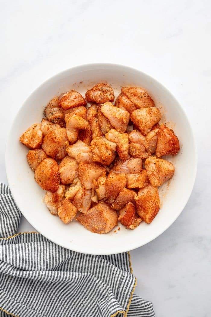 Overhead view of raw bite-sized chicken breast pieces in a white bowl after being tossed with Cajun-style seasonings. The chicken is evenly coated in a reddish-orange spice mixture. The bowl sits on a light gray marble surface beside a gray-and-white striped kitchen towel with yellow trim.