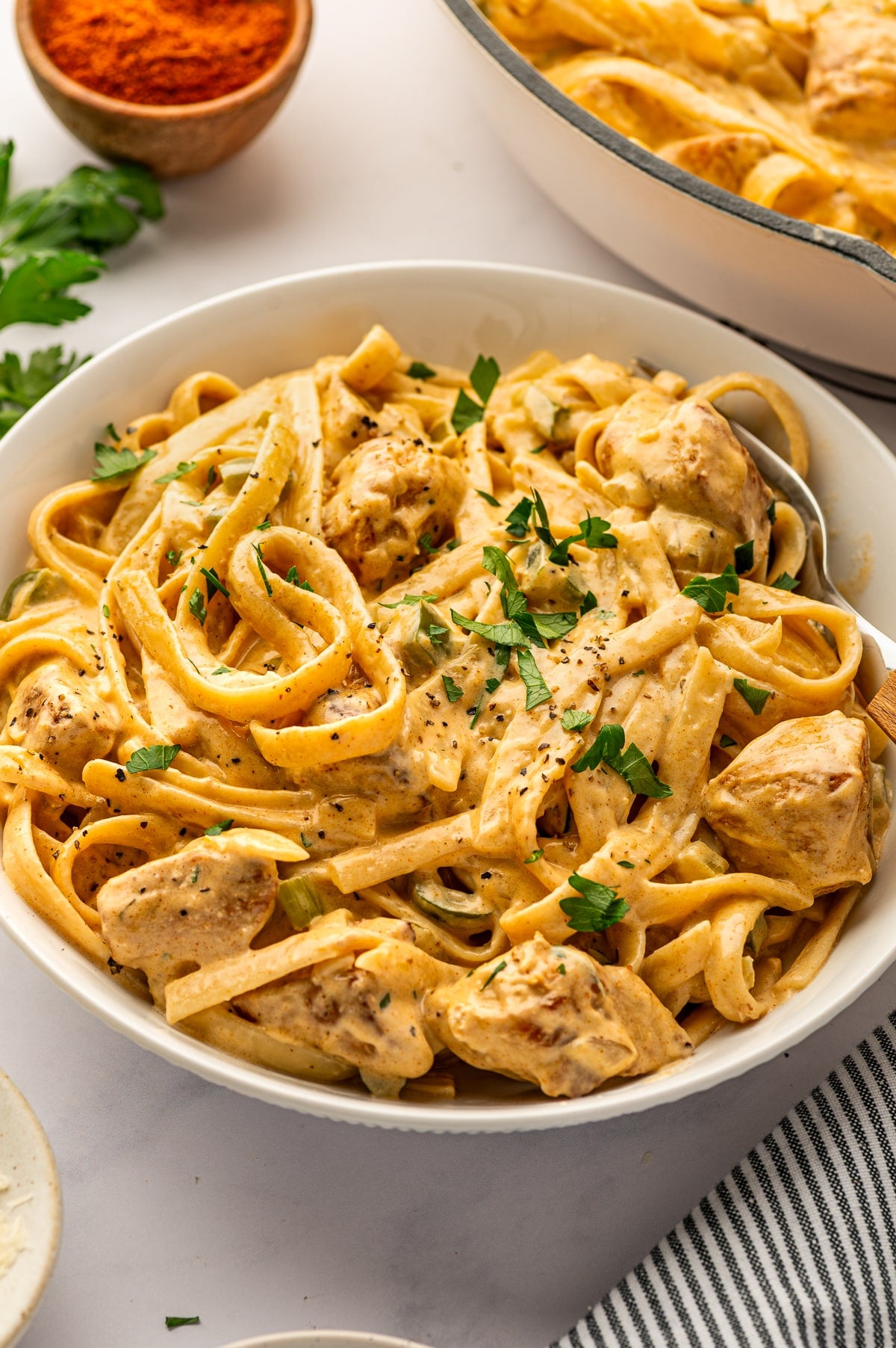 Overhead angled view of Cajun Chicken Alfredo served in a large white bowl with a silver spoon. The bowl is filled with creamy fettuccine, browned bite-sized chicken pieces, chopped parsley, and black pepper. In the background, part of the cream-colored skillet is visible along with a small wooden bowl of Cajun seasoning, fresh parsley, and a gray-and-white striped kitchen towel.