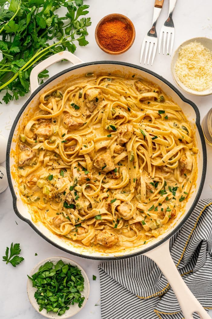 Overhead view of finished Cajun Chicken Alfredo in a cream-colored skillet with a black rim and pale handle. Fettuccine noodles are coated in creamy orange Cajun Alfredo sauce and mixed with bite-sized chicken pieces, chopped parsley, and black pepper. Around the skillet are fresh parsley sprigs, a wooden bowl of Cajun seasoning, a bowl of grated Parmesan, a bowl of chopped parsley, two forks with wooden handles, a small bottle of olive oil, and a gray-and-white striped kitchen towel with yellow trim on a light gray marble surface.