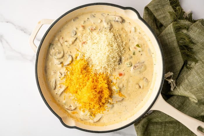 White enameled skillet filled with creamy mushroom sauce topped with two piles of cheese, bright orange shredded cheddar and finely grated white parmesan, photographed overhead on a white marble counter with a green towel to the right.
