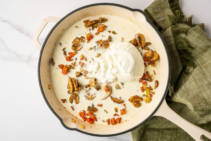 Overhead view of a white enameled skillet with broth and cream poured over sautéed vegetables and mushrooms, with a large dollop of sour cream sitting in the center and a green linen towel beside the pan.