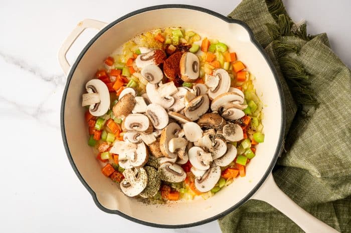 Overhead view of a white enameled skillet filled with sautéed carrots, celery, and onion topped with sliced mushrooms, a mound of smoked paprika, dried Italian seasoning, and cracked black pepper, with a green linen towel to the side.