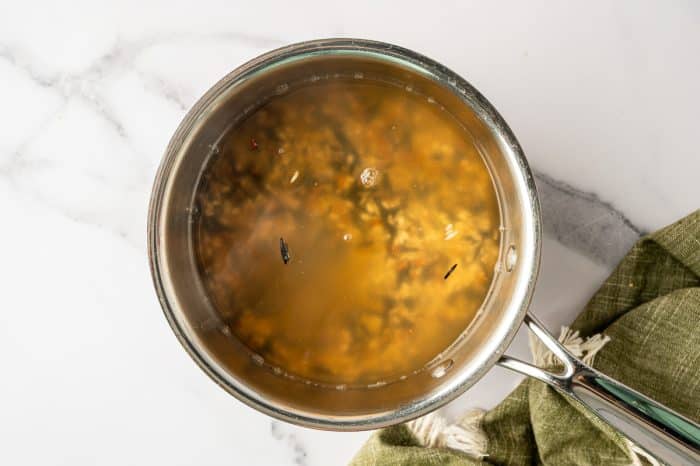 Overhead view of a stainless steel saucepan filled with golden chicken broth and wild rice blend simmering, set on a white marble countertop with a green linen towel on the right.