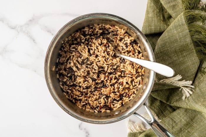 Cooked wild rice blend in a stainless steel saucepan with a metal spoon resting inside, photographed from above on a white marble counter with a green towel in the background. Cooked wild rice blend in a stainless steel saucepan with a metal spoon resting inside, photographed from above on a white marble counter with a green towel in the background.