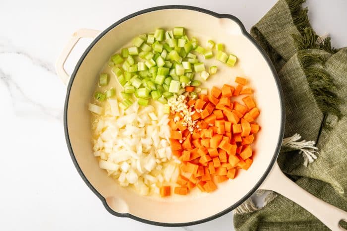 Overhead view of a white enameled skillet with butter melting around diced celery, chopped onion, diced carrots, and minced garlic, on a white marble surface with a green cloth napkin beside the pan.