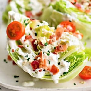 Large white rectangular serving platter with four wedges of iceberg lettuce, cut-side-up, drizzled with homemade blue cheese dressing and sprinkled with crumbled bacon bits, halved cherry tomatoes, chives, and blue cheese crumbles.