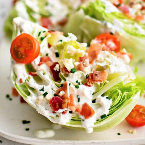 Large white rectangular serving platter with four wedges of iceberg lettuce, cut-side-up, drizzled with homemade blue cheese dressing and sprinkled with crumbled bacon bits, halved cherry tomatoes, chives, and blue cheese crumbles.