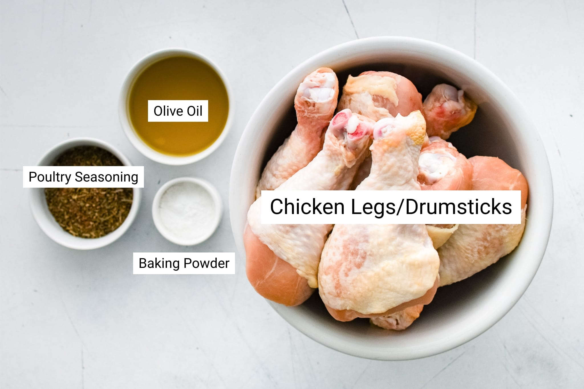 Overhead view of raw chicken drumsticks in a white bowl surrounded by small bowls of olive oil, poultry seasoning, and baking powder, each labeled.