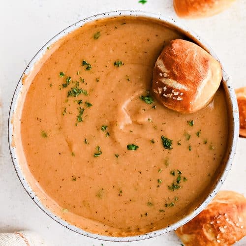 One small, soft pretzel bite resting in a large serving bowl filled with beer cheese dip garnished with fresh parsley