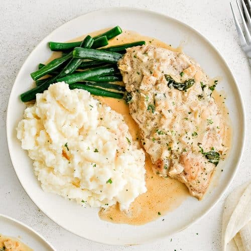 White serving plate filled with one piece of cooked chicken, a scoop of mashed potatoes, and pan-fried green beans all smothered in creamy boursin cheese sauce.