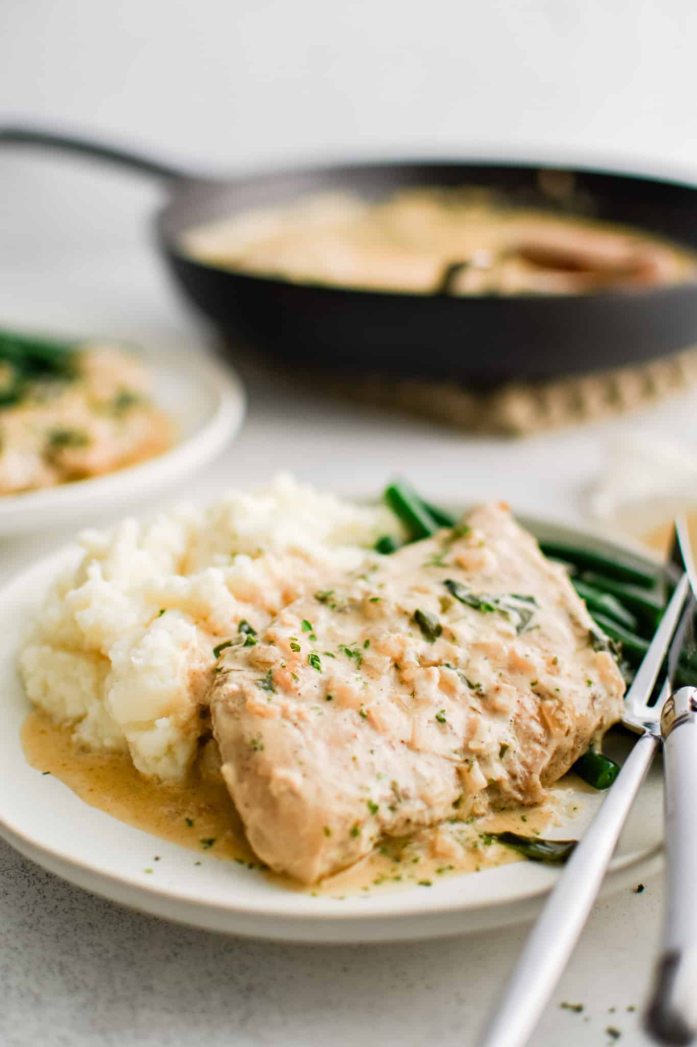 White serving plate filled with one piece of cooked chicken, a scoop of mashed potatoes, and pan-fried green beans all smothered in creamy boursin cheese sauce.
