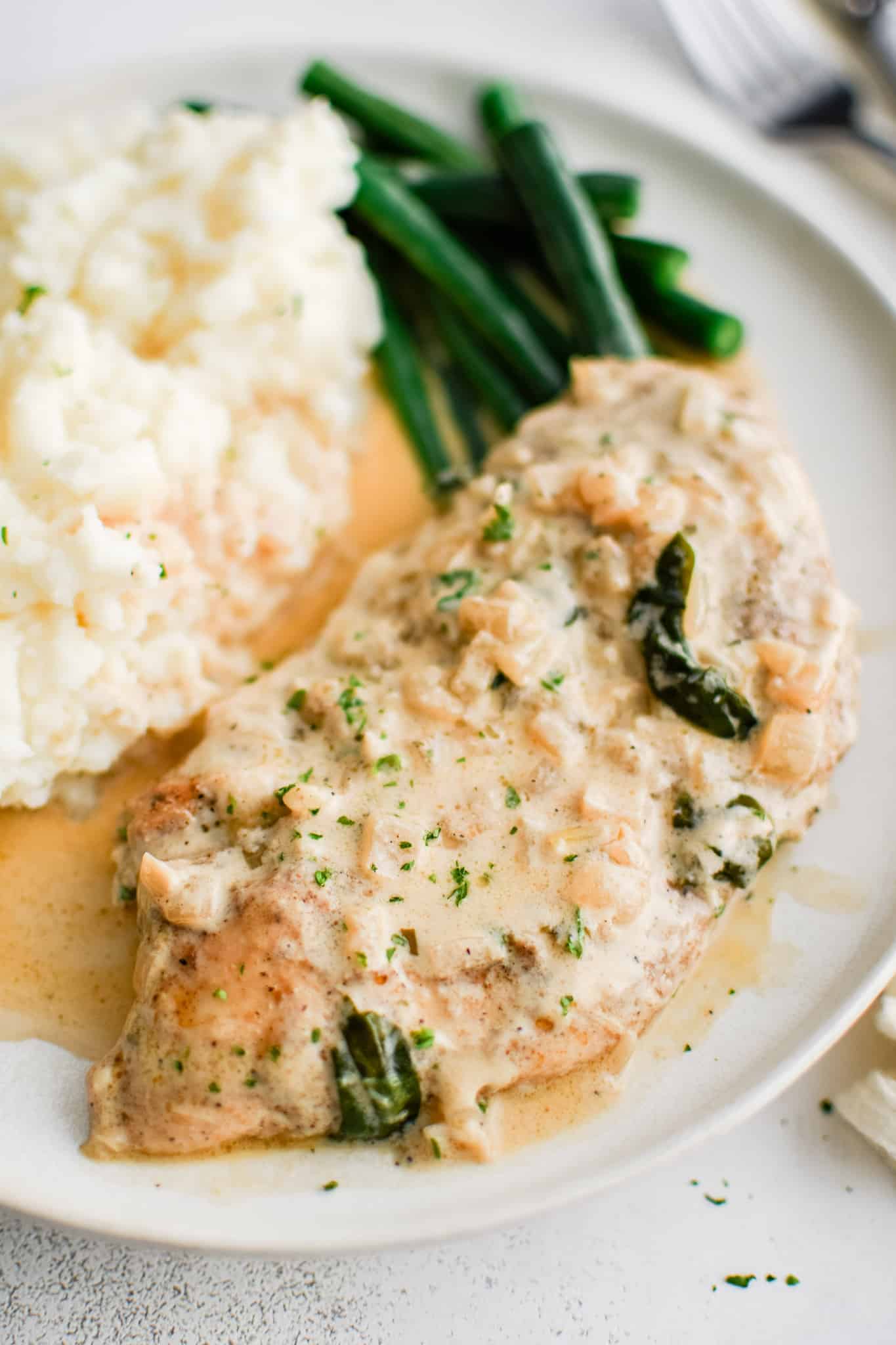White serving plate filled with one piece of cooked chicken, a scoop of mashed potatoes, and pan-fried green beans all smothered in creamy boursin cheese sauce.