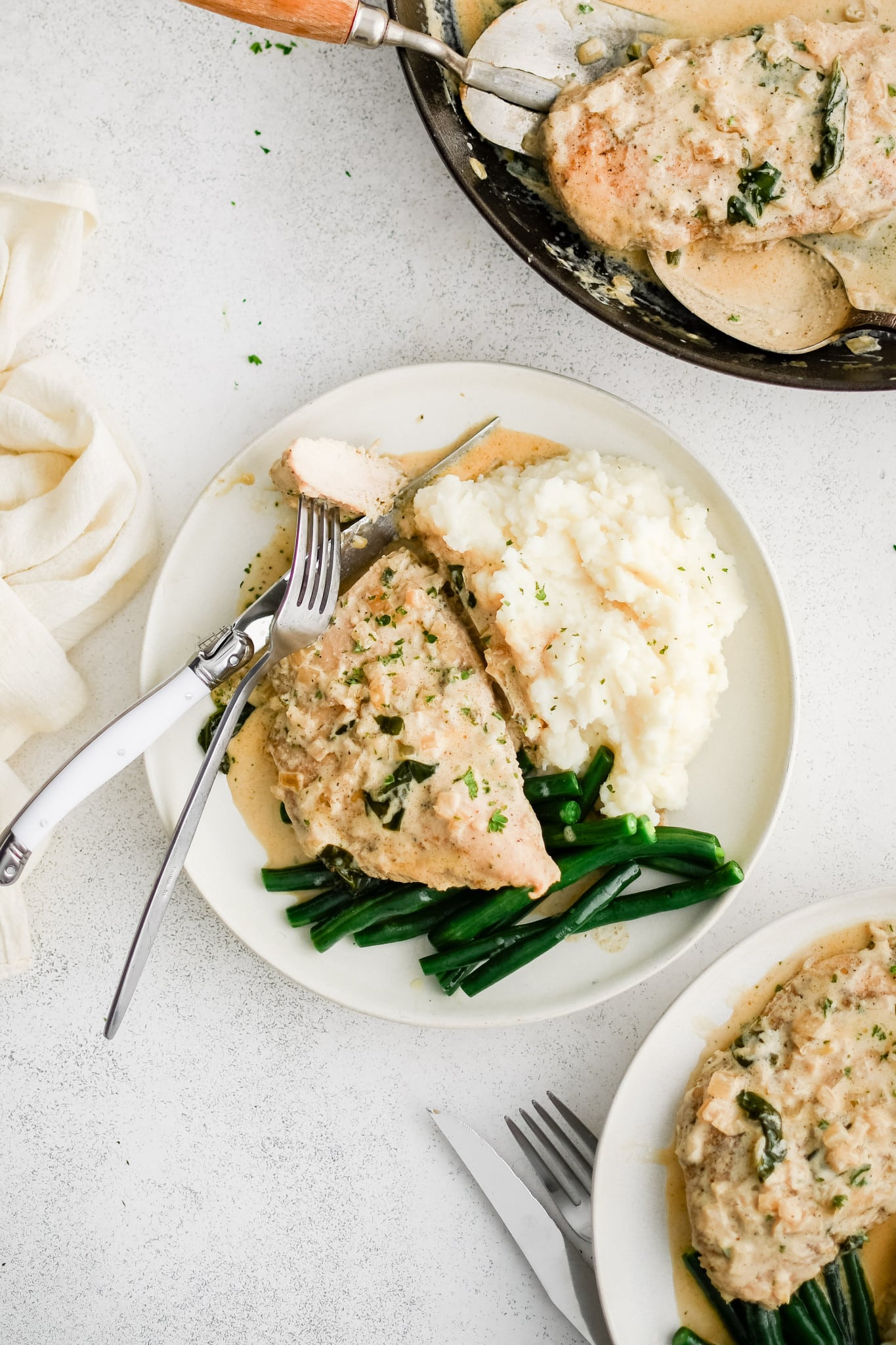 Three white serving plate filled with one piece of cooked chicken, a scoop of mashed potatoes, and pan-fried green beans all smothered in creamy boursin cheese sauce.