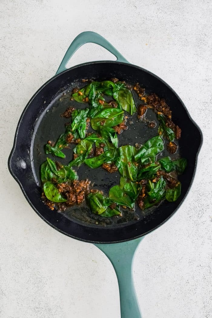 Large skillet filled with wilted spinach, sun dried tomatoes, and minced garlic cooking in butter.