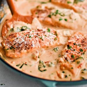 Large pan filled with five cooked salmon fillets resting in a creamy sauce made with heavy cream, sun dried tomatoes, spinach, garlic, and parmesan cheese and garnished with fresh parsley and more parmesan cheese.