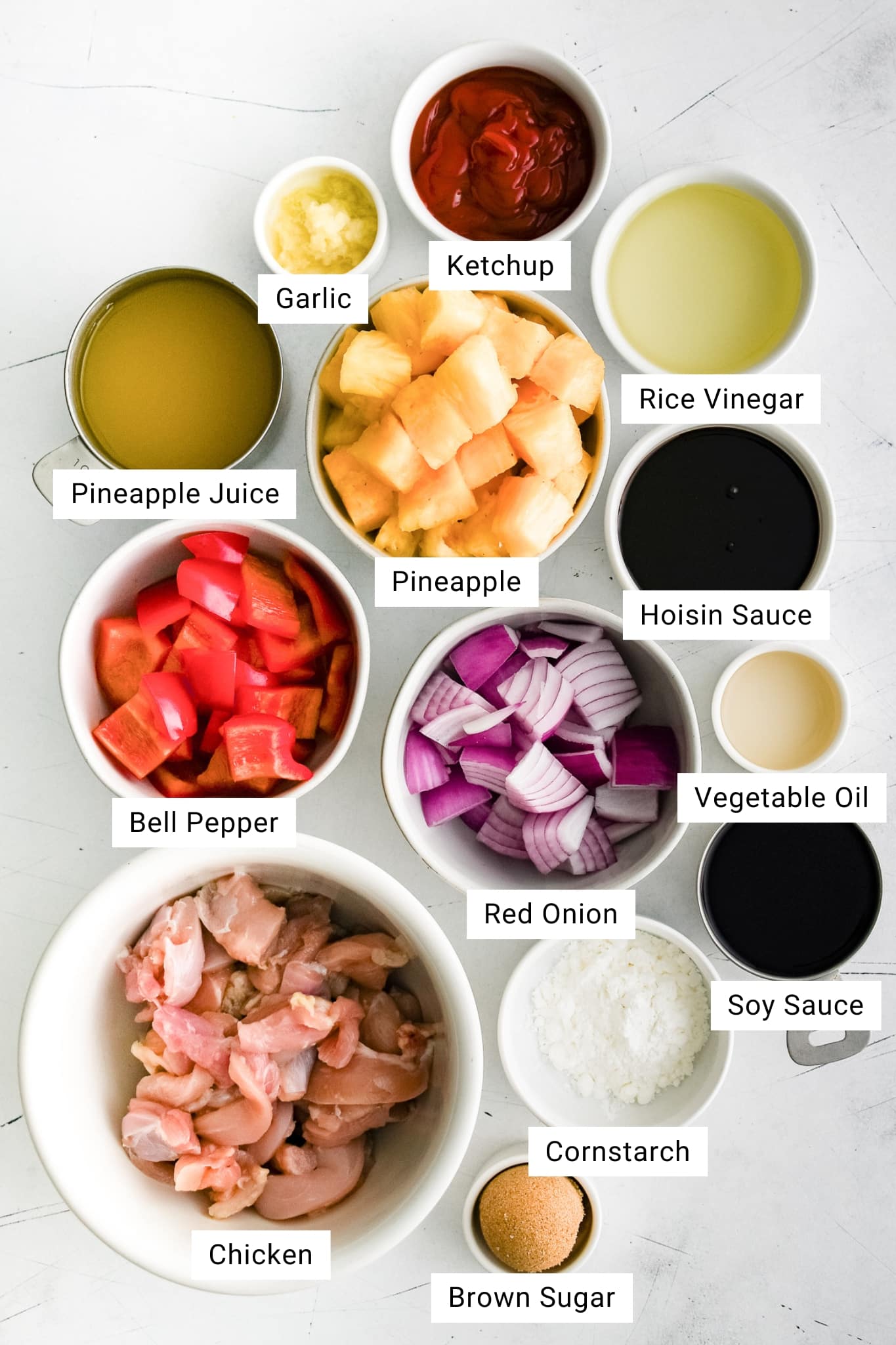 Overhead view of all measured ingredients for pineapple chicken in small bowls, including chicken, pineapple juice, hoisin sauce, soy sauce, rice vinegar, ketchup, garlic, onion, bell pepper, cornstarch, and brown sugar.